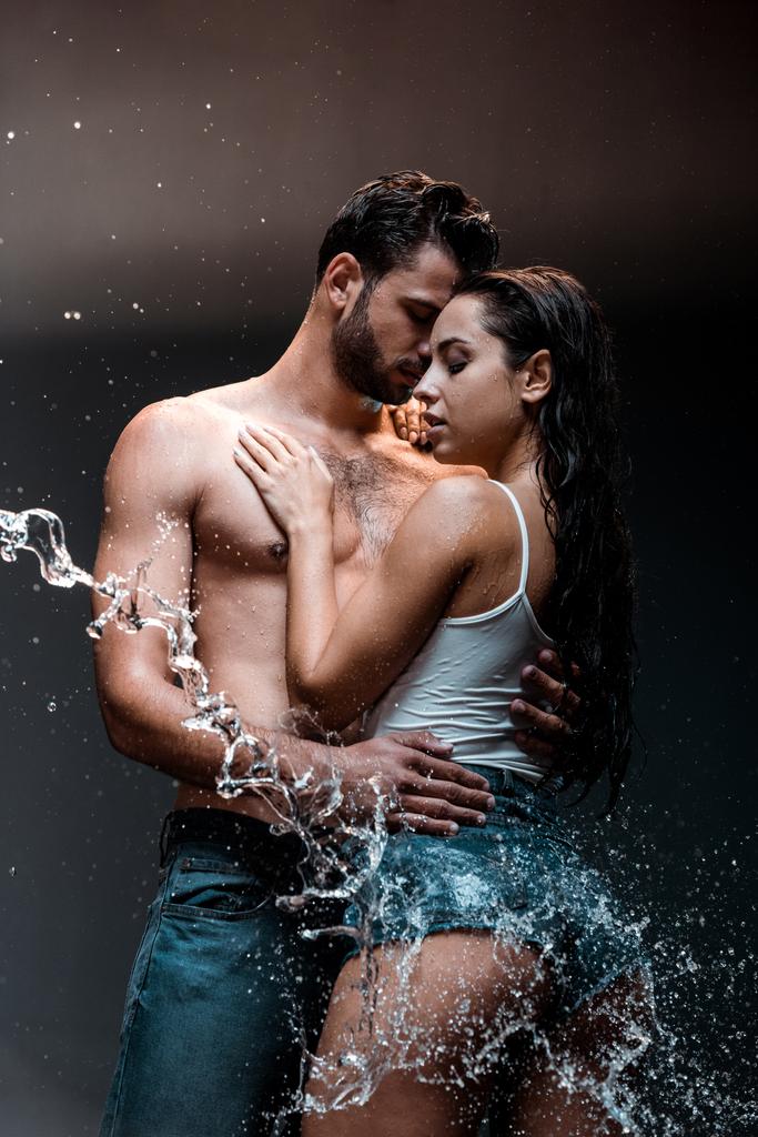 uomo bello e senza maglietta abbracciando fidanzata vicino spruzzata d'acqua sul nero
  - Foto, immagini