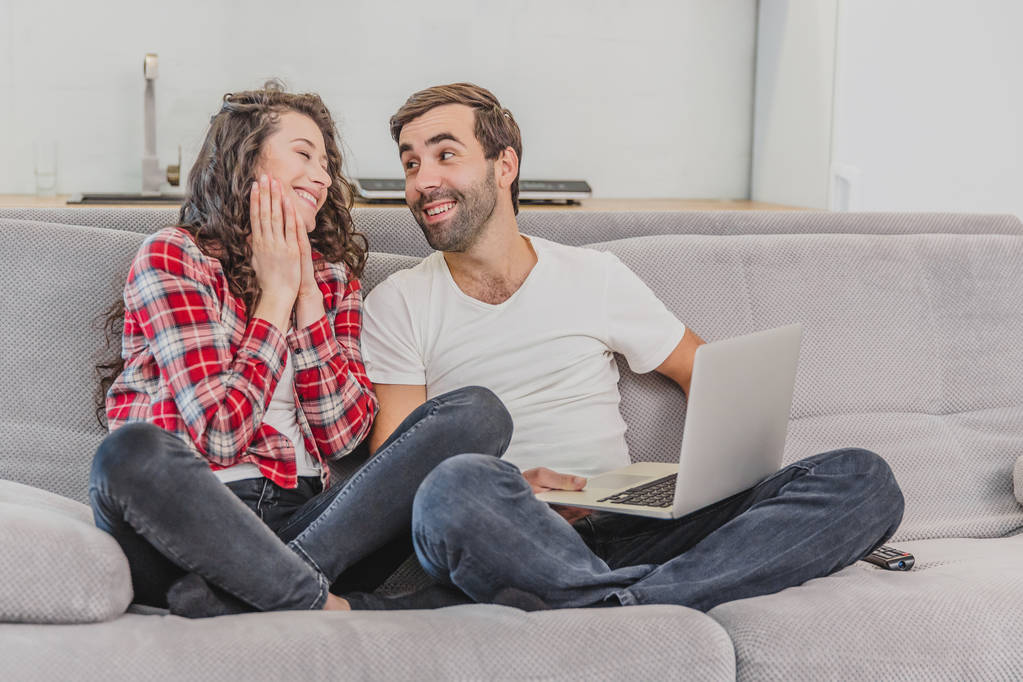 Молодая пара весело проводит время за компьютером, сидя на диване дома вместе, счастливые мужчина и женщина весело смотрят на экран ноутбука
. - Фото, изображение