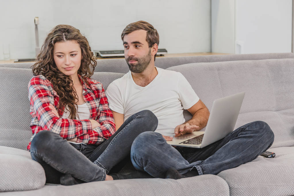 Хорошая пара во время покупок в Интернете с помощью ноутбука и улыбаясь, сидя на диване дома. Женщина была оскорблена мужчиной, потому что он не сделал ей покупки в Интернете
. - Фото, изображение