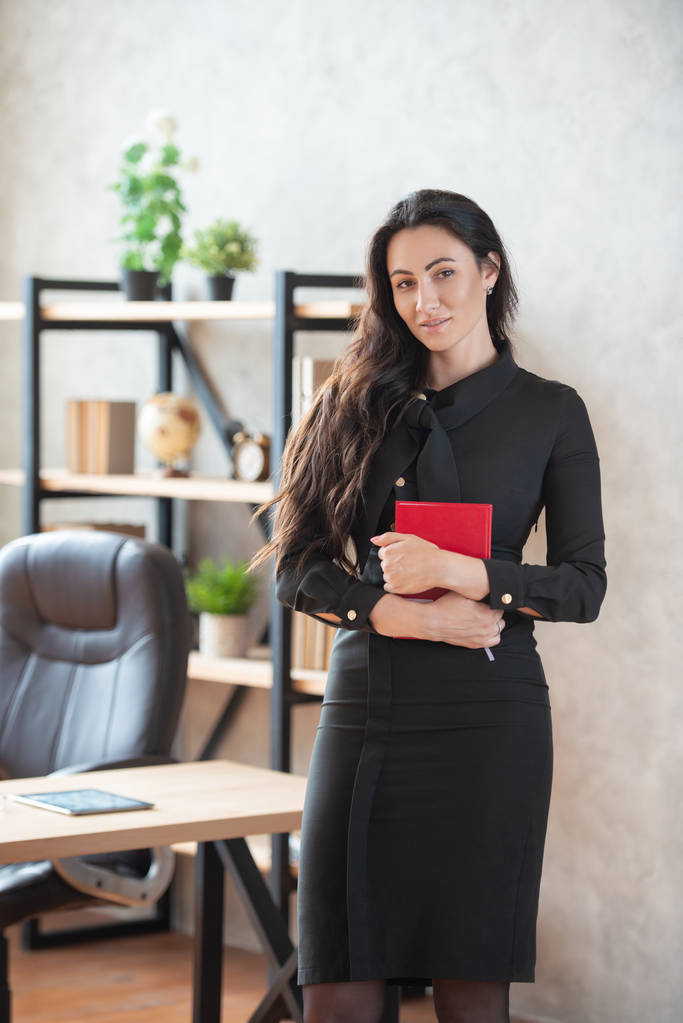 Mulher de negócios elegante bonita com um caderno vermelho na mão está de pé no escritório. Negócios e carreira das mulheres
 - Foto, Imagem