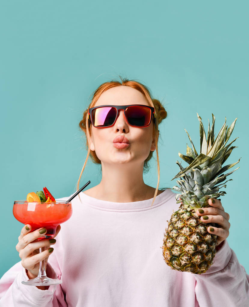 Joyful young woman in modern sunglasses with tropic cocktail strawberry margarita and pineapple in her hands send us a kiss - Photo, Image