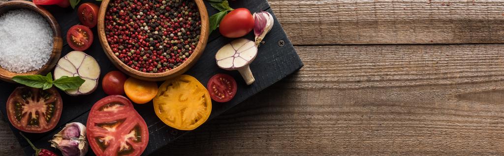 panoramatický záběr černého pultu s mísy s pepřem a solí v blízkosti chilli papriček, špenátu, rajčat nakrájené na plátky a česneku na dřevěném stole  - Fotografie, Obrázek