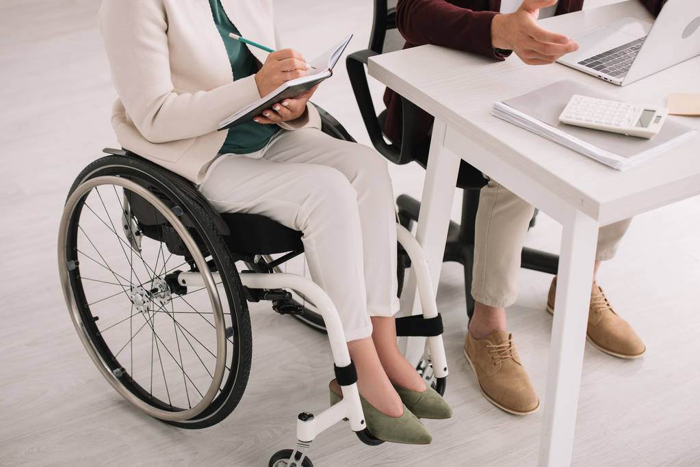 bijgesneden beeld van gehandicapte zakenvrouw schrijven in notitieboekje tijdens het zitten in een rolstoel in de buurt van collega - Foto, afbeelding