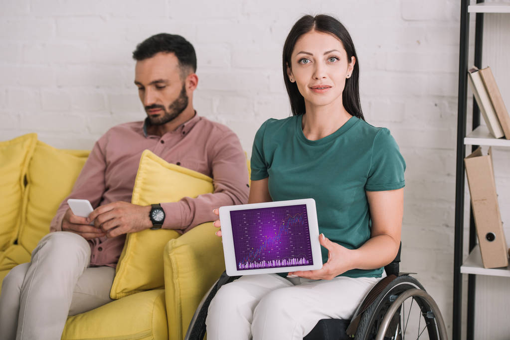 Lächelnde behinderte Frau zeigt digitales Tablet mit Infografik auf dem Bildschirm, während sie neben ihrem Freund sitzt und das Smartphone benutzt - Foto, Bild