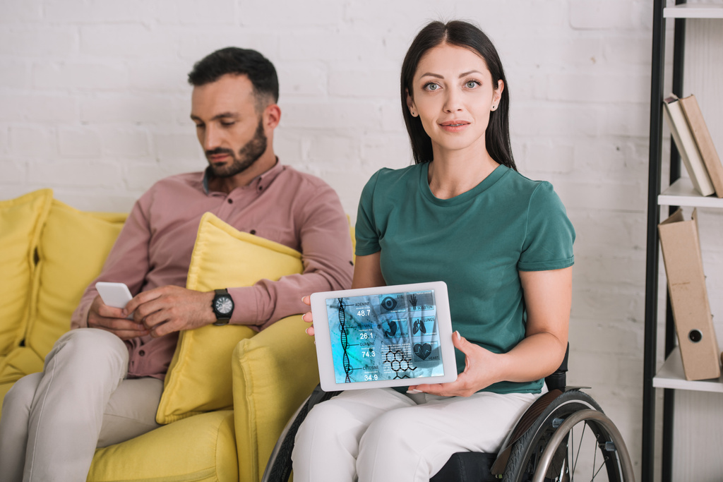 jonge gehandicapte vrouw toont digitale tablet met DNA-informatie app terwijl zittend in de buurt van vriendje met behulp van smartphone - Foto, afbeelding