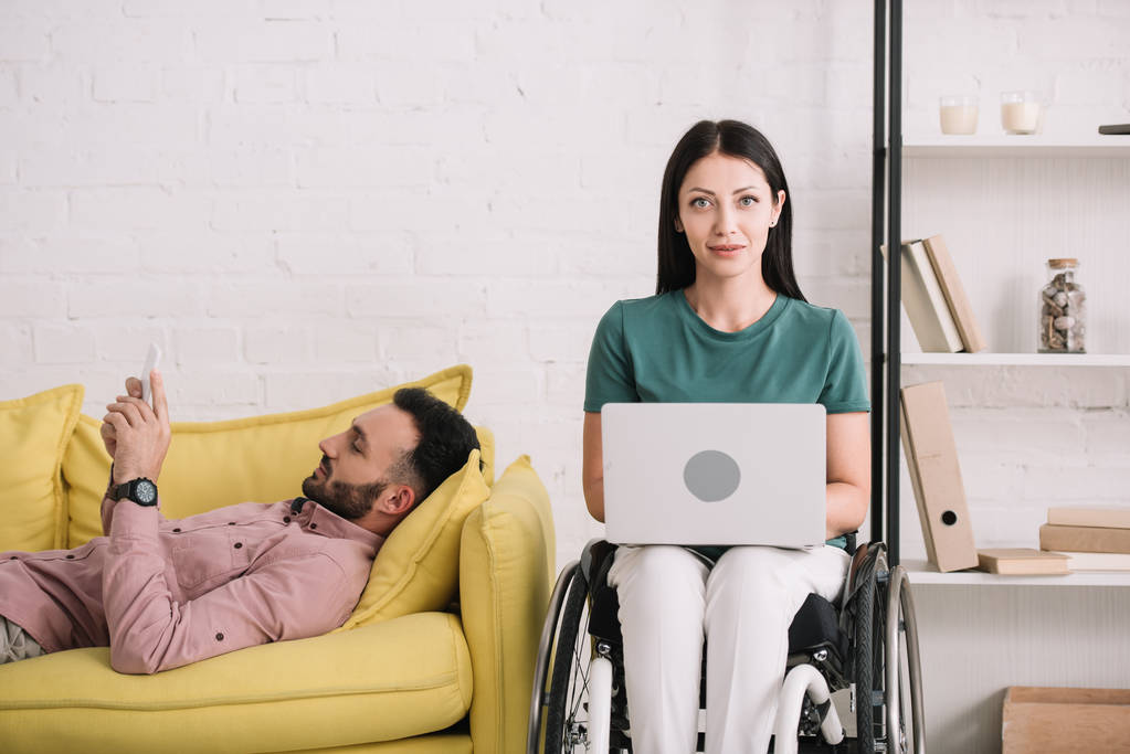 улыбающаяся женщина-инвалид с помощью ноутбука, сидя рядом с парнем, лежащим на диване и используя смартфон
 - Фото, изображение