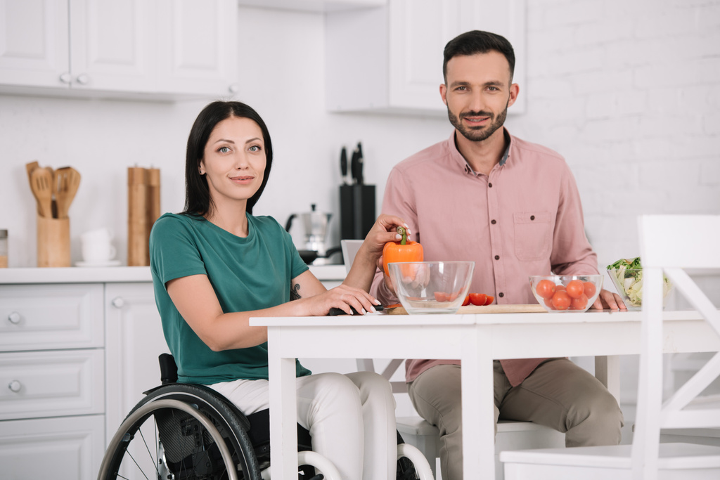 привлекательная женщина-инвалид с парнем, улыбающимся в камеру, сидя за кухонным столом и готовящим салат вместе
 - Фото, изображение
