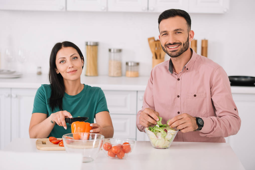щаслива пара дивиться на камеру, сидячи за кухонним столом і готуючи салат разом
 - Фото, зображення
