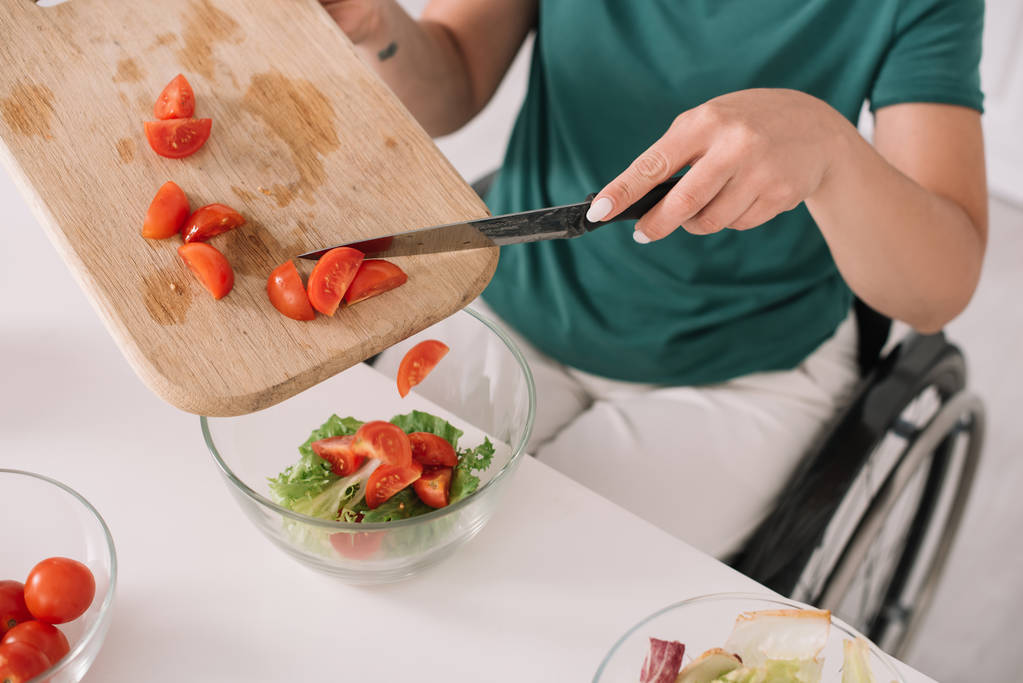 vista parcial de la mujer discapacitada la adición de tomates en rodajas en tazón de vidrio en la cocina
 - Foto, imagen