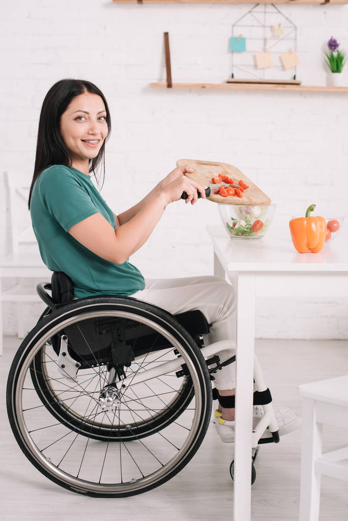 vrolijke gehandicapte vrouw glimlachend bij de camera tijdens het bereiden van salade in de keuken - Foto, afbeelding