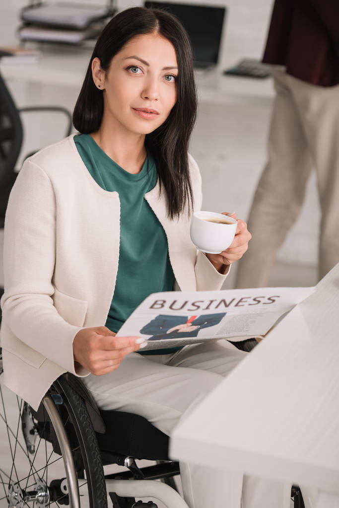 atraente mulher de negócios deficiente segurando xícara de café e jornal enquanto olha para a câmera
 - Foto, Imagem