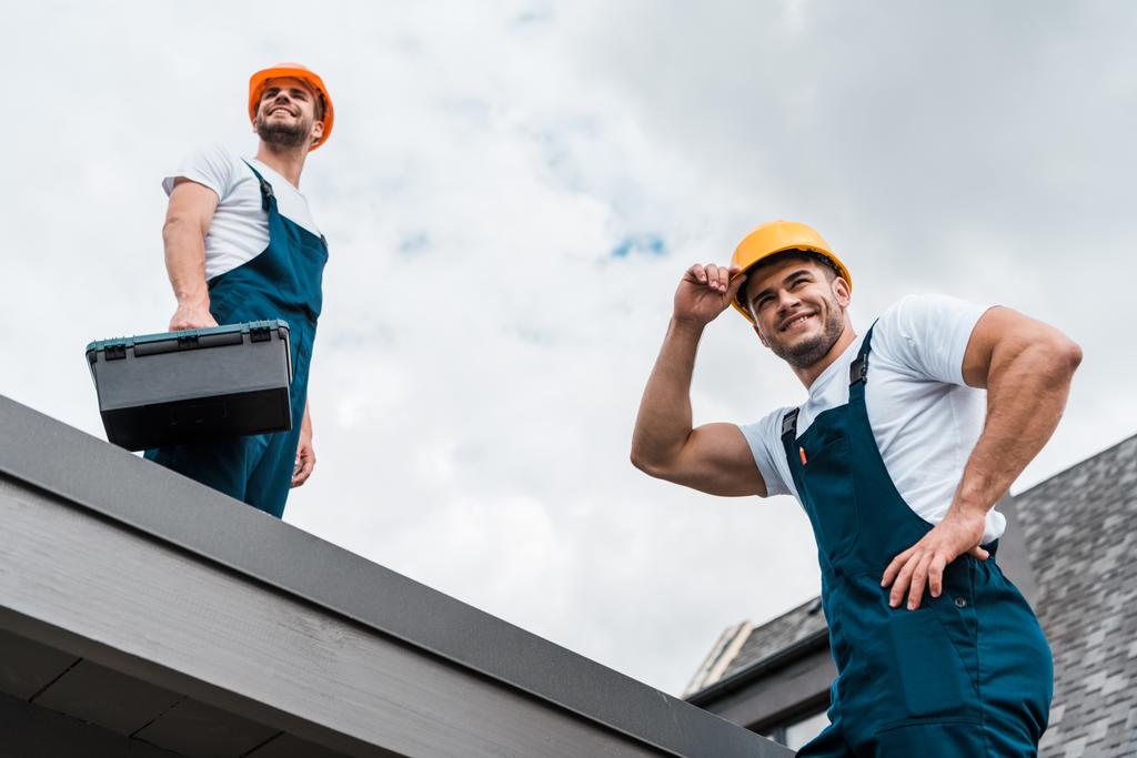 Tiefansicht glücklicher Heimwerker in Helmen gegen wolkenverhangenen Himmel  - Foto, Bild