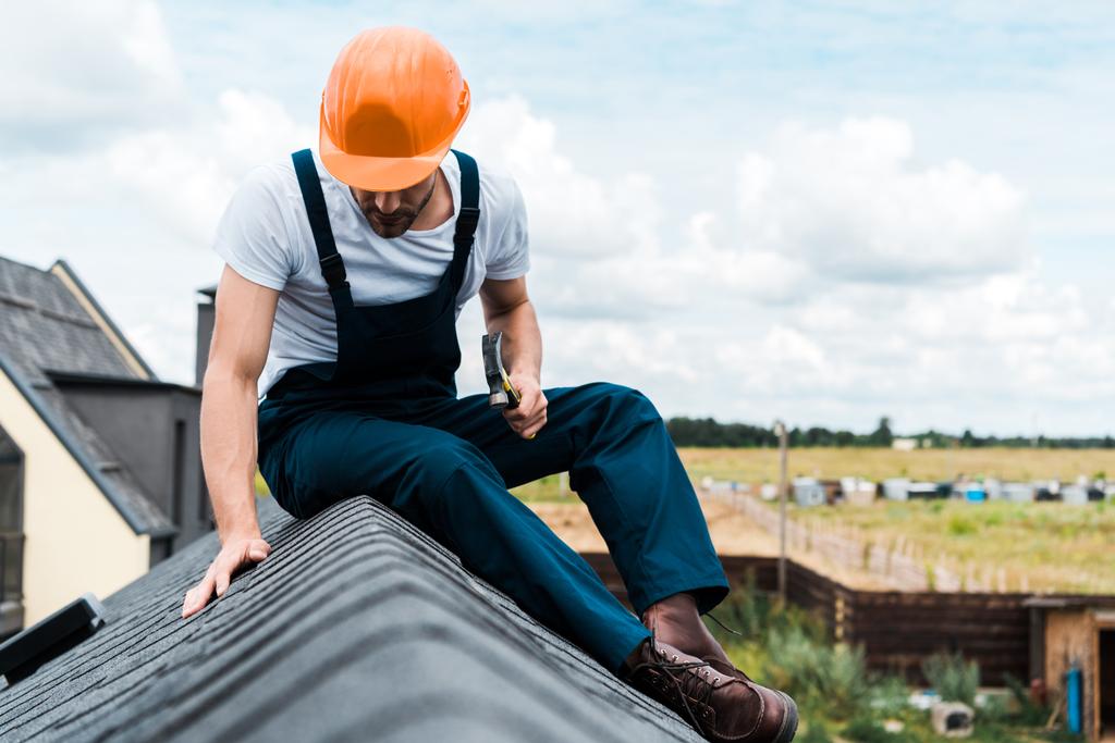 enfoque selectivo del manitas en casco naranja sentado en el techo y sosteniendo el martillo
  - Foto, Imagen