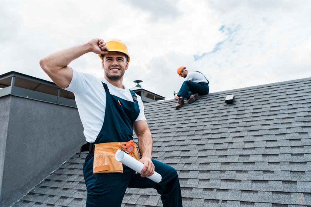 Selektiver Fokus glücklicher Reparateure, die Rollenpapier halten, während Mitarbeiter Dach reparieren  - Foto, Bild