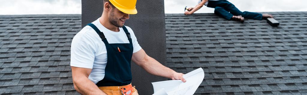 панорамный снимок красивого строителя, держащего чертеж, пока коллега чинит крышу
  - Фото, изображение