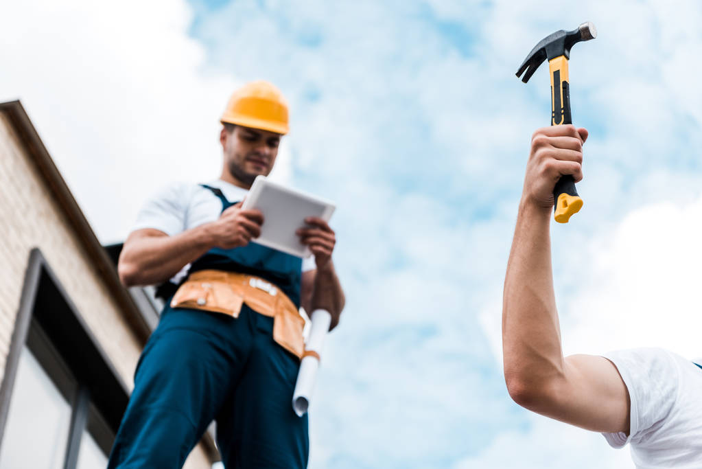 selective focus of man holding hammer near coworker in helmet using digital tablet  - Photo, Image