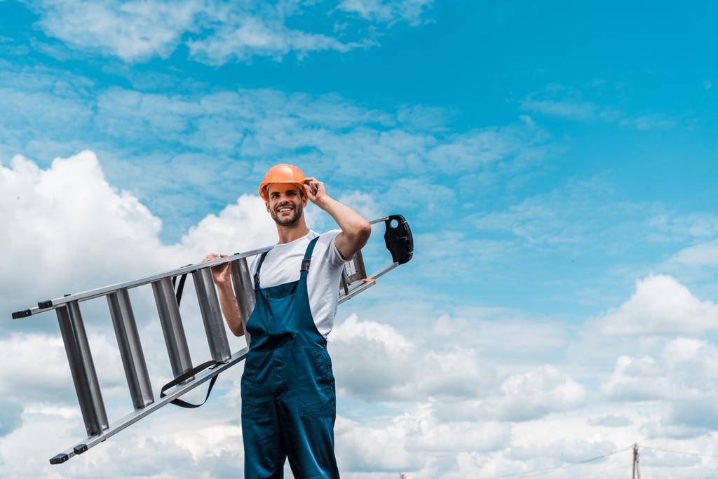 χαρούμενος επισκευαστής κρατώντας σκάλα και χαμογελά κατά του γαλάζιου ουρανού με σύννεφα  - Φωτογραφία, εικόνα