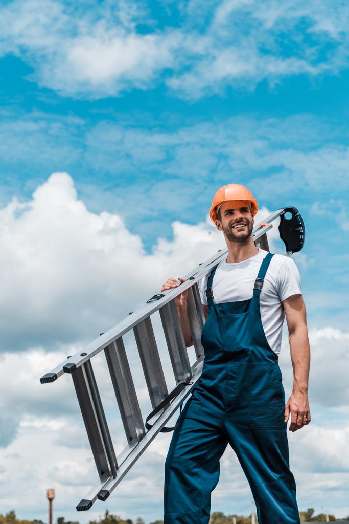 reparador feliz sosteniendo escalera y sonriendo contra el cielo azul con nubes
  - Foto, Imagen