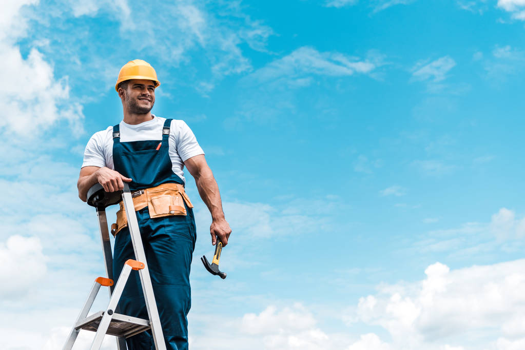happy repairman standing on ladder and smiling against blue sky with clouds  - Photo, Image