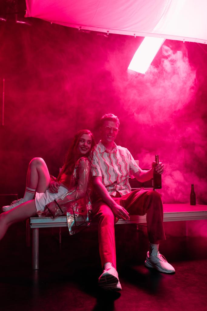 Χαμογελαστός άνθρωπος με μπύρα κάθεται κοντά κορίτσι σε νυχτερινό κλαμπ κατά τη διάρκεια του ρέιβ - Φωτογραφία, εικόνα