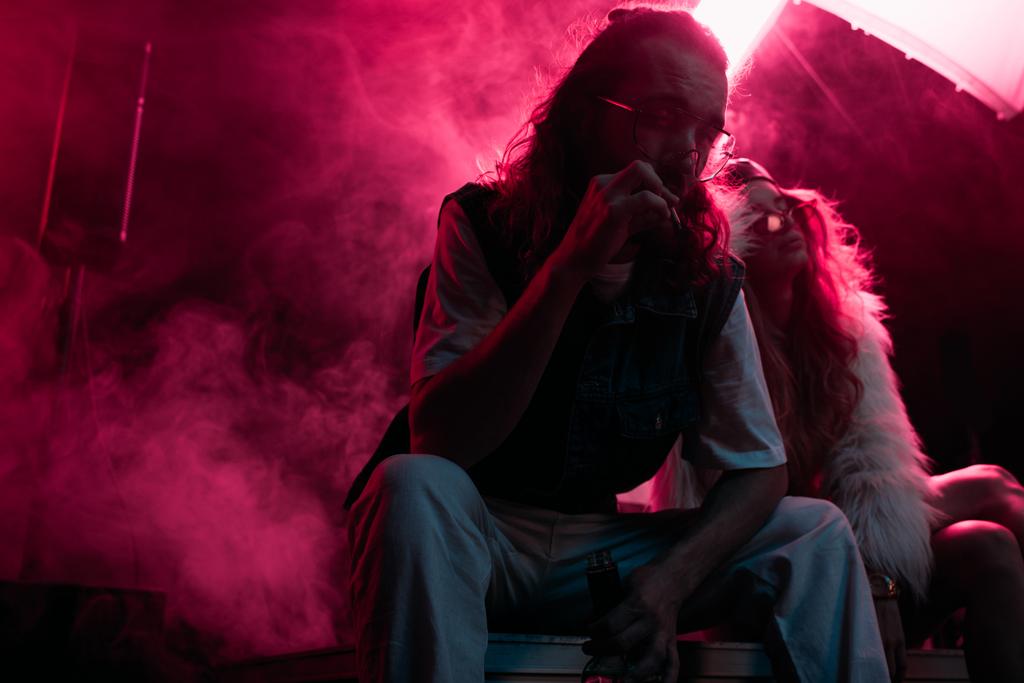 mężczyzna palenia papierosów w pobliżu młodej kobiety podczas rave party w klubie nocnym - Zdjęcie, obraz