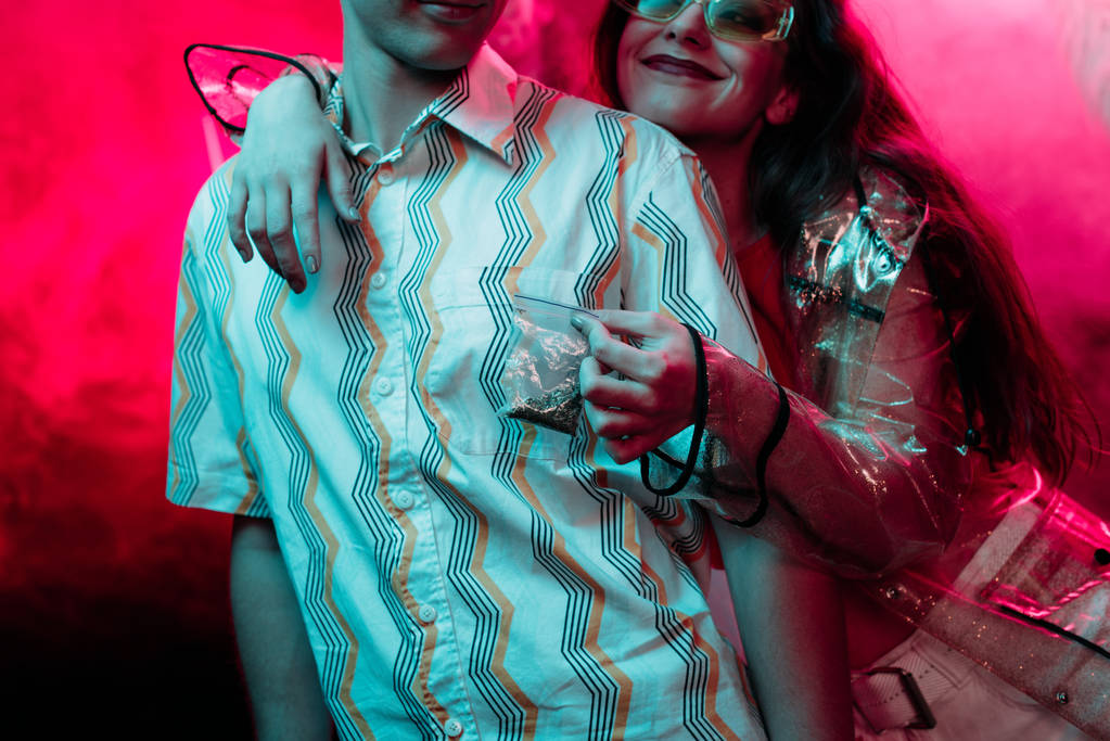 Обрезанный вид девушки, держащей травку в пластиковом пакете молнии рядом с мужчиной в ночном клубе
 - Фото, изображение