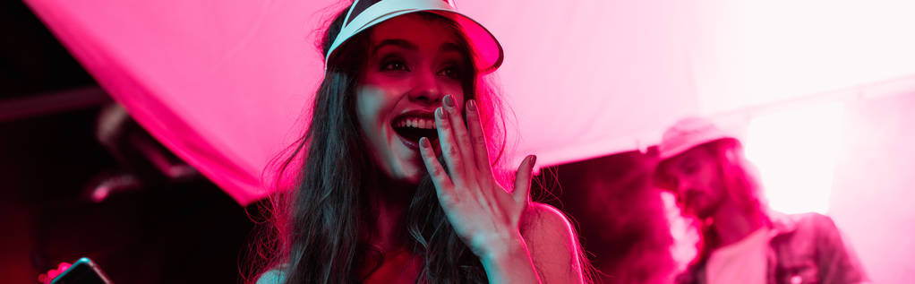 Panoramaaufnahme eines aufgeregten Mädchens, das während einer Rave-Party in einem Nachtclub den Mund bedeckt  - Foto, Bild