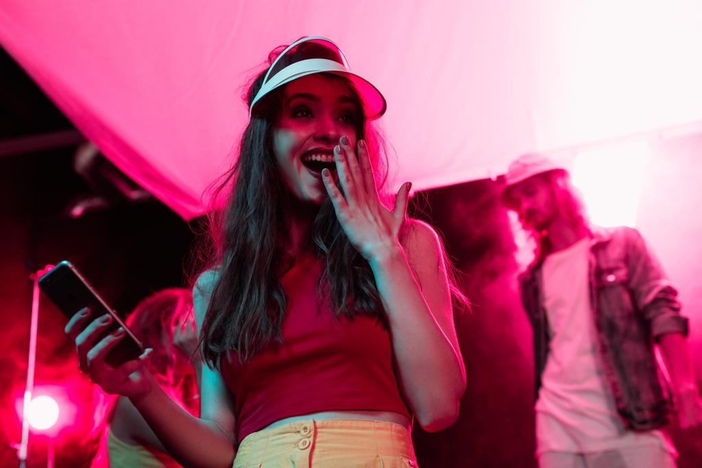 έκπληκτος κορίτσι με το smartphone που καλύπτει το στόμα κατά τη διάρκεια ρέιβ πάρτι σε νυχτερινό κέντρο - Φωτογραφία, εικόνα