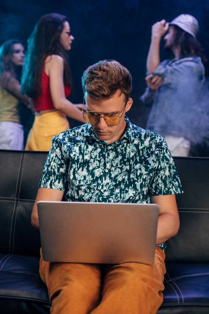hombre guapo en gafas de sol sentado en el sofá y el uso de ordenador portátil en el club nocturno
 - Foto, imagen