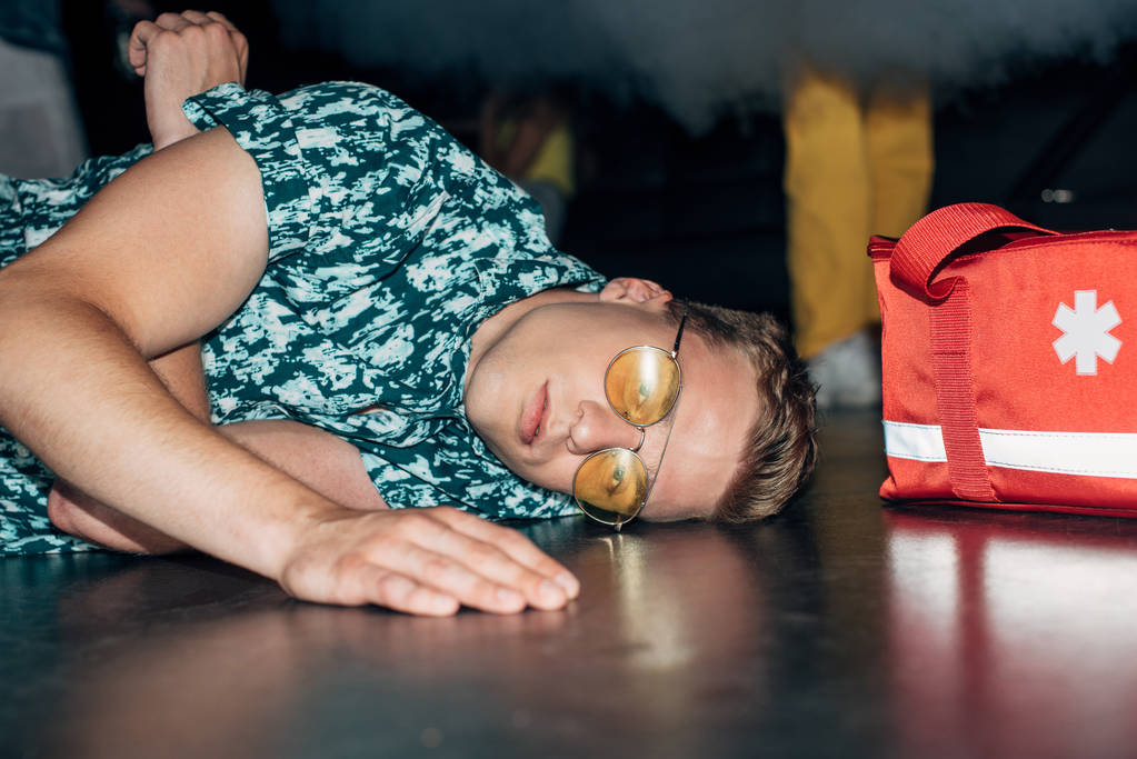 хворий чоловік в сонцезахисних окулярах лежить на підлозі біля аптечки для першої допомоги в нічному клубі
 - Фото, зображення