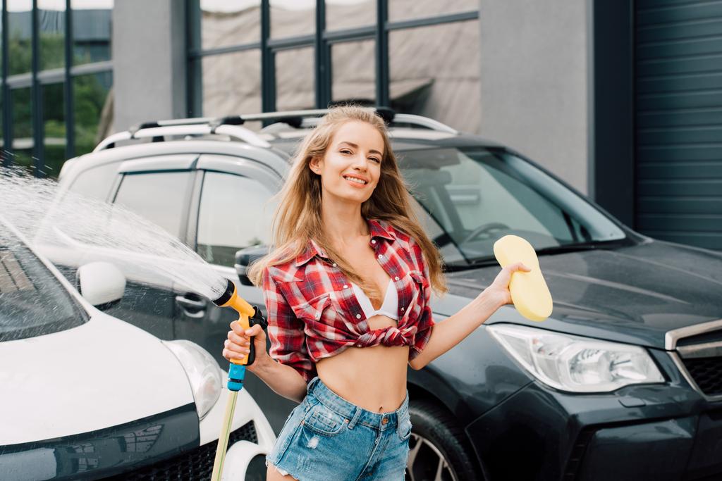alegre chica sosteniendo presión lavadora y esponja cerca de coches
  - Foto, Imagen