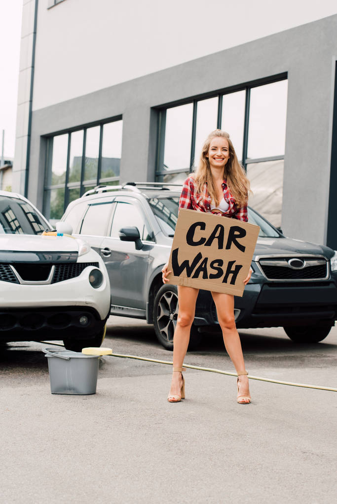 pozytywna dziewczyna stoi i trzyma karton z myjni samochodowej w pobliżu samochodów  - Zdjęcie, obraz
