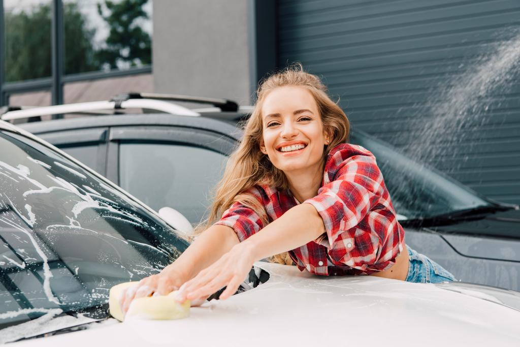 mulher positiva lavando para-brisa do carro com esponja e espuma
  - Foto, Imagem