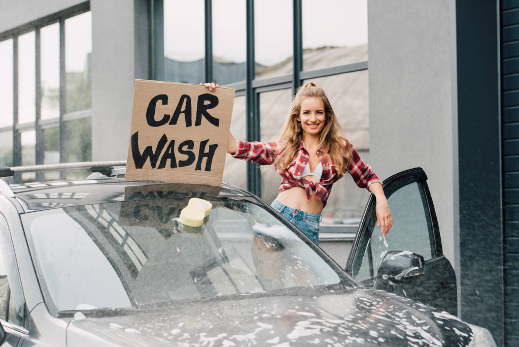 pozytywna młoda kobieta trzymania kartonu z napisem myjni samochodowej i stoi w pobliżu samochodów  - Zdjęcie, obraz