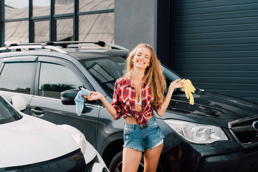 vrolijke vrouw houdt blauwe doek en gele latex handschoenen in de buurt van auto's   - Foto, afbeelding