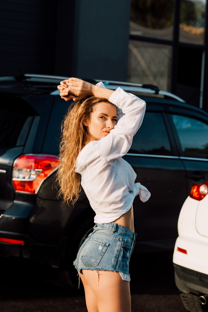 atraente jovem mulher em calções jeans azuis olhando para a câmera perto de carros
  - Foto, Imagem