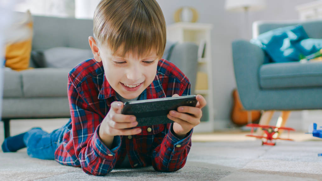 Inteligentne Little Boy układanie na dywan odgrywa w grze wideo na jego smartphone, trzyma telefon komórkowy w poziomym trybie krajobrazu. Dziecko ma zabawa gra Videogame w Sunny Living Pokój. - Zdjęcie, obraz