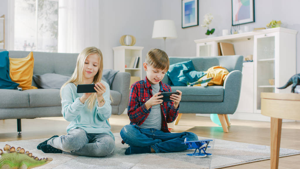 W domu siedzi na dywanie: Cute Little Girl i Sweet Boy grając w konkurencyjnej gry wideo na dwóch smartfonach, trzymając je w poziomym trybie krajobrazu. - Zdjęcie, obraz