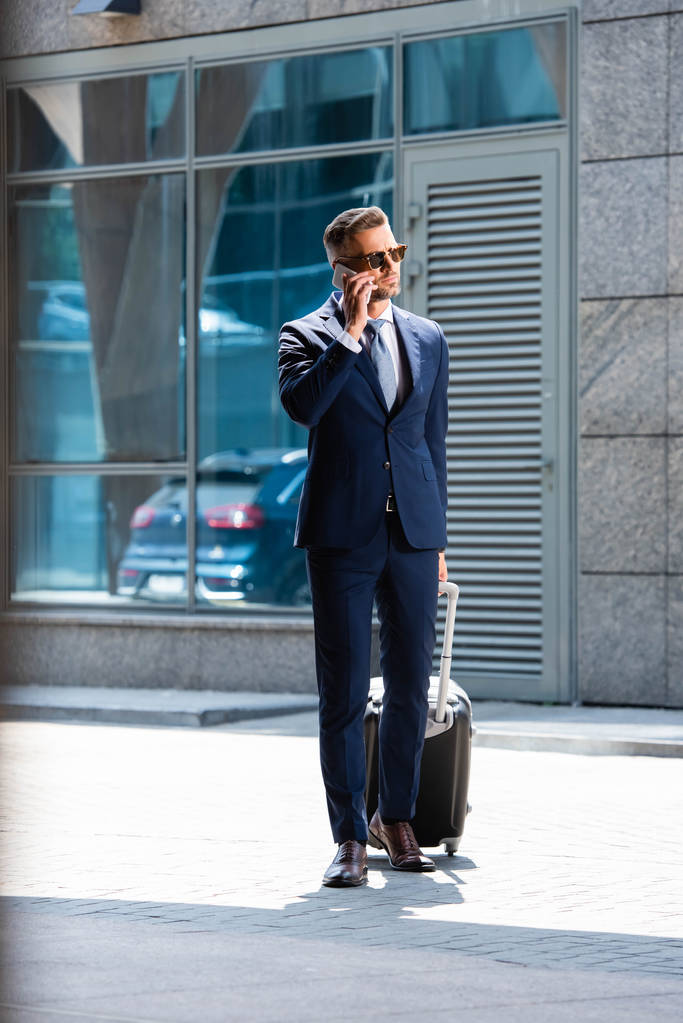 όμορφος άντρας με κουστούμι και γυαλιά κρατώντας βαλίτσα και μιλώντας στο smartphone  - Φωτογραφία, εικόνα