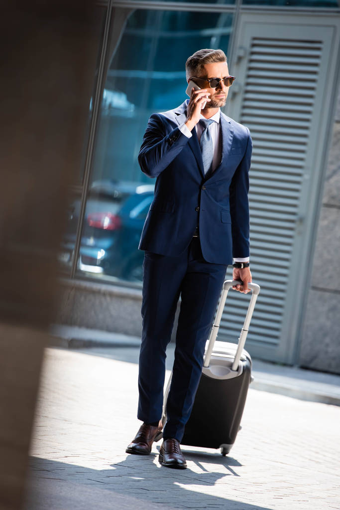 スマートフォンで話し、スーツケースを保持するスーツとメガネでハンサムなビジネスマンの選択的な焦点 - 写真・画像
