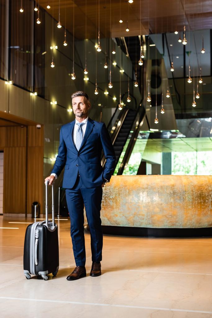 schöner Geschäftsmann im Anzug mit der Hand in der Tasche, die Koffer hält - Foto, Bild