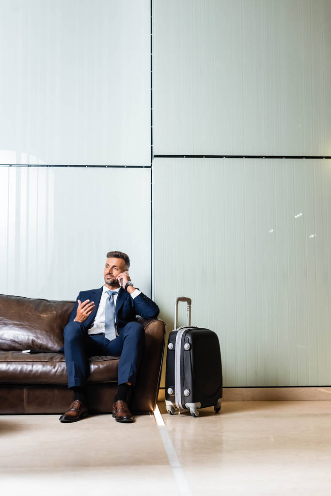 schöner Geschäftsmann im Anzug sitzt auf dem Sofa und spricht auf dem Smartphone  - Foto, Bild
