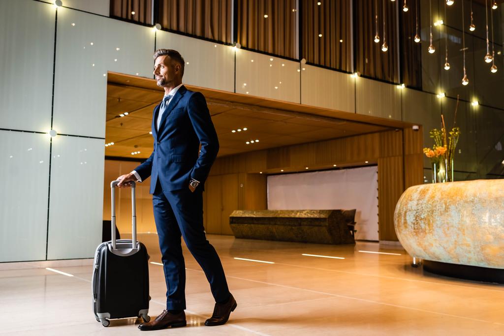 hombre de negocios guapo en traje con la mano en el bolsillo que sostiene la maleta
 - Foto, Imagen