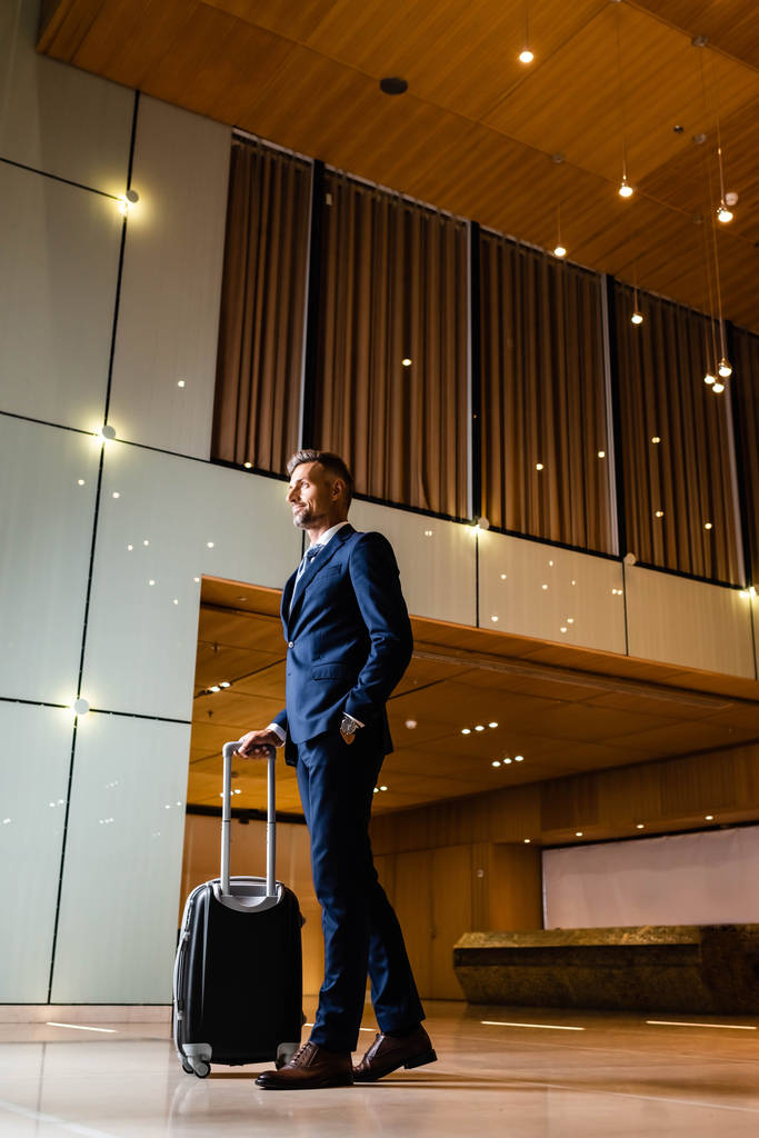 hombre de negocios guapo en traje con la mano en el bolsillo que sostiene la maleta
 - Foto, Imagen