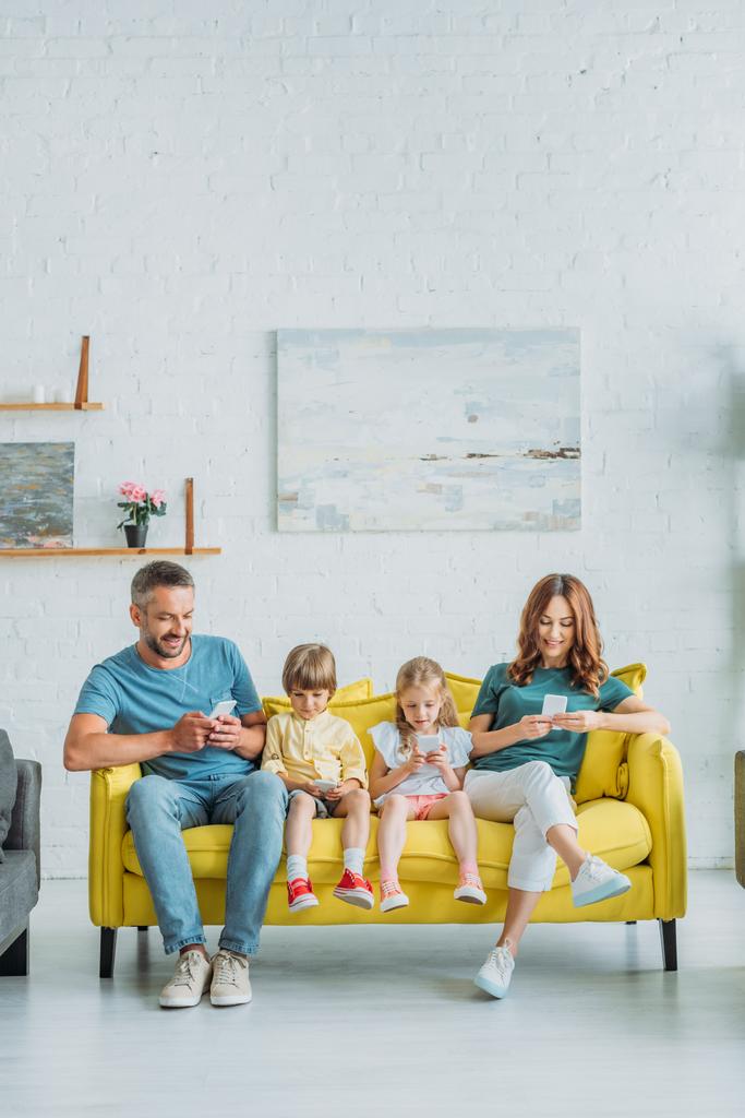 自宅で黄色いソファに座っている間、スマートフォンを使用して家族を笑顔  - 写真・画像