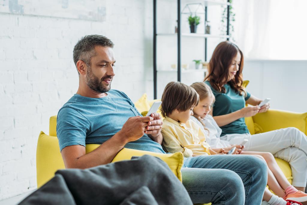 happy parents with daughter and son using smartphones while sitting on sofa at home - Photo, Image