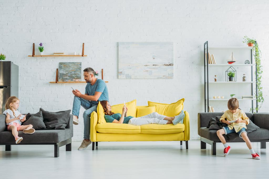 mãe e pai com dois filhos usando smartphones enquanto descansam em sofás em casa
 - Foto, Imagem