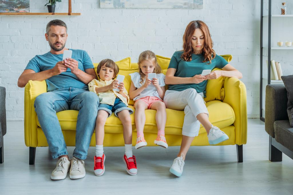 προσεκτική οικογένεια που κάθεται σε κίτρινο καναπέ στο σπίτι και χρησιμοποιώντας smartphones - Φωτογραφία, εικόνα