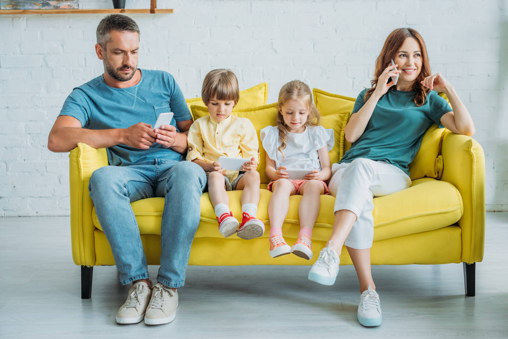 Lächelnde Frau, die mit dem Smartphone spricht, während sie neben Mann und Kindern auf der Couch sitzt und Smartphones benutzt  - Foto, Bild