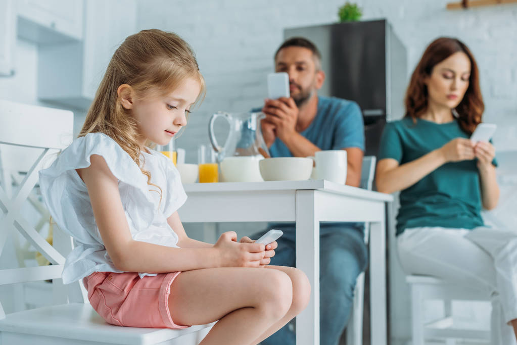 selective focus of child with smartphone sitting at table with served breakfast near parents using smartphones - Photo, Image